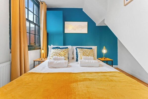 A bedroom with a bed, two bedside tables, a window with curtains, a wall-mounted artwork, and a sloped ceiling.