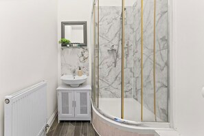Une salle de bain avec un lavabo blanc, un miroir, une douche avec une porte en verre et un mur en marbre.
