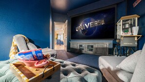 A home theater room with a large flat-screen TV displaying 'UNIVERSAL', a reclining chair, a coffee table with snacks, and a vintage Nostalgia popcorn machine. - Welcome to the Coco Cabana! Located in Estrella Mountain Ranch in Goodyear, AZ! (Goodyear)