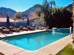 A swimming pool with lounge chairs and umbrellas, surrounded by a landscaped garden and mountains in the background.