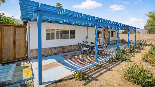 Un patio con una pérgola azul, una mesa con sillas y una alfombra estampada.
