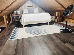A cozy attic bedroom with a bed, a bedside table, a lamp, a fan, and a rug.
