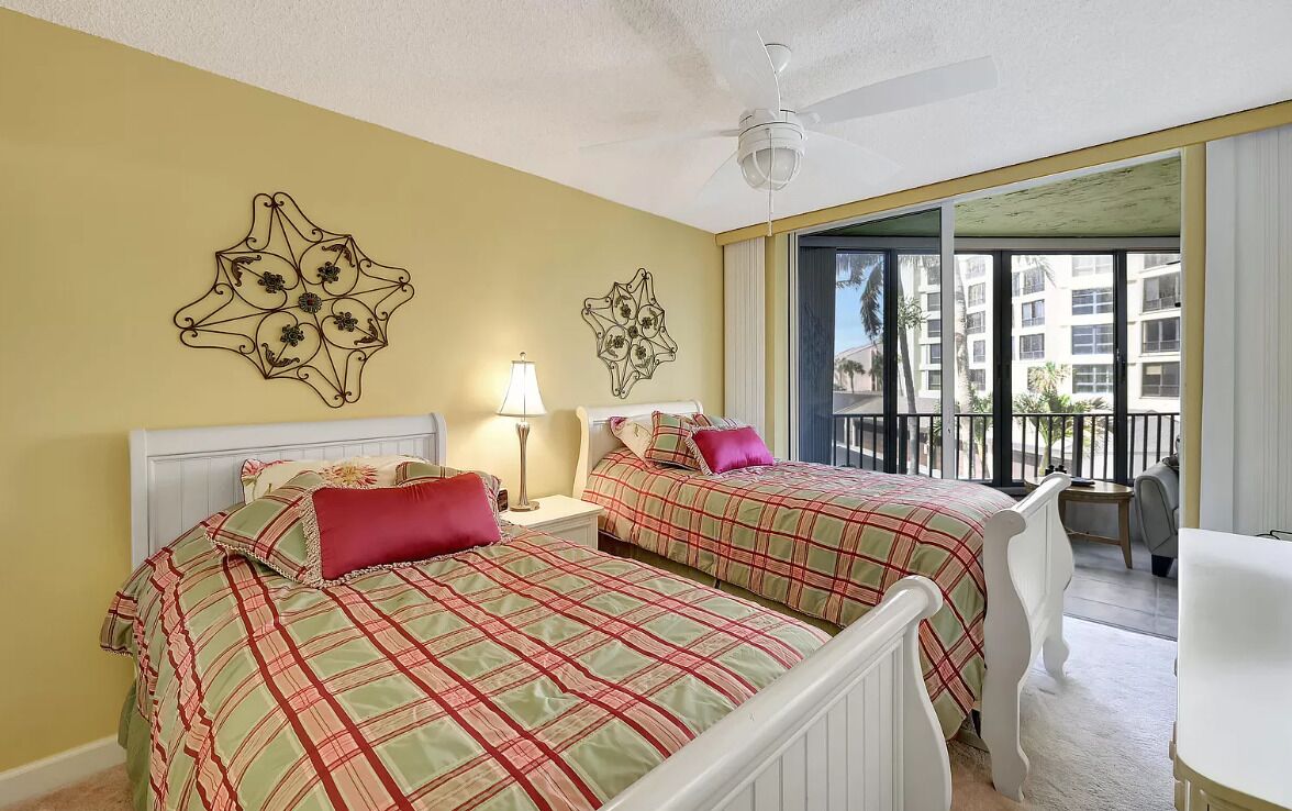 A hotel room with two beds, a ceiling fan, a balcony with a view of buildings, and a chair.