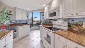 A kitchen with white cabinets, granite countertops, and a view of palm trees through large windows.