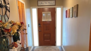 A hallway with a wooden floor, a dark wooden console table with a vase and a basket, a door with a 'Bless This Home' sign, and a light fixture above.
