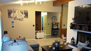 A living room with a grey sofa, a coffee table, a lamp, and a TV mounted on a white brick wall. There is a clock on the wall and a door leading to another room.