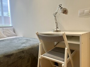 A small, foldable desk with a desk lamp, a grey cushioned chair, and a bed with a grey blanket in a minimalistic room.