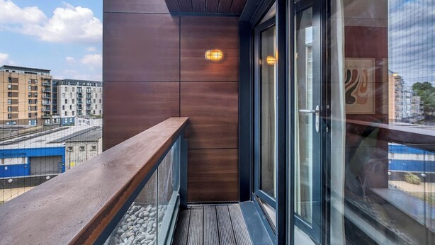 A balcony with a wooden railing, a glass door, and a view of surrounding buildings. - Modern 1BR • 2 min to Tube • O2 & ExCeL (London)