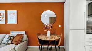 A modern living room with a sofa, a round wooden dining table with chairs, a white cabinet, and a round mirror on the wall. - Modern 1BR • 2 min to Tube • O2 & ExCeL (London)