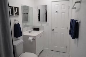 Un baño con inodoro, lavamanos y espejo. Hay toallas colgadas en la pared y una puerta.