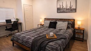 A bedroom with a bed, two bedside lamps, a desk with a laptop, a chair, and a cactus painting on the wall.