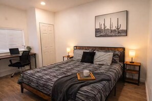 Un dormitorio con cama, dos lámparas de noche, un escritorio con una computadora portátil, una silla y una pintura de un cactus en la pared.