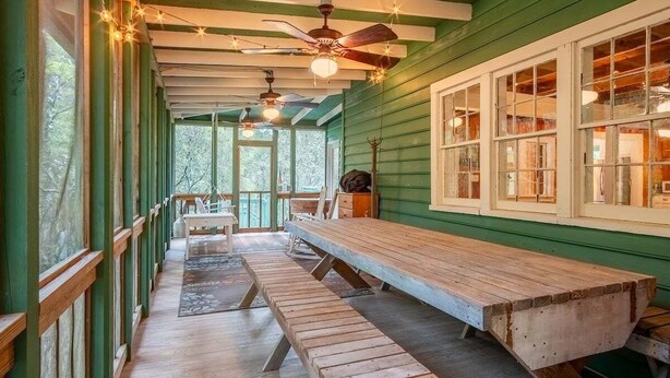 A covered outdoor dining area with wooden benches and tables, a ceiling fan, and large windows.