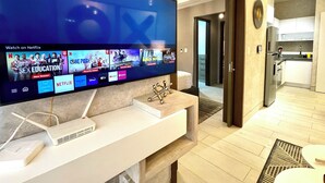 A modern hotel room with a flat-screen TV displaying Netflix content, a white desk with a laptop, and a view into the kitchen area.