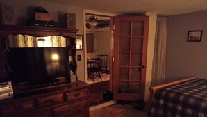 Une pièce faiblement éclairée, avec une commode en bois, un téléviseur, un tapis à motifs et un lit recouvert d’une couverture à carreaux.