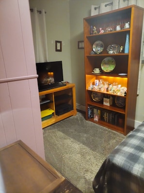 A compact living space with a television on a wooden stand, a bookshelf with various items, and a small table with a yellow container.