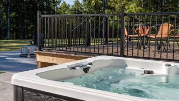 Un jacuzzi con revestimiento de madera, rodeado por una valla de metal, con mobiliario exterior y árboles al fondo.