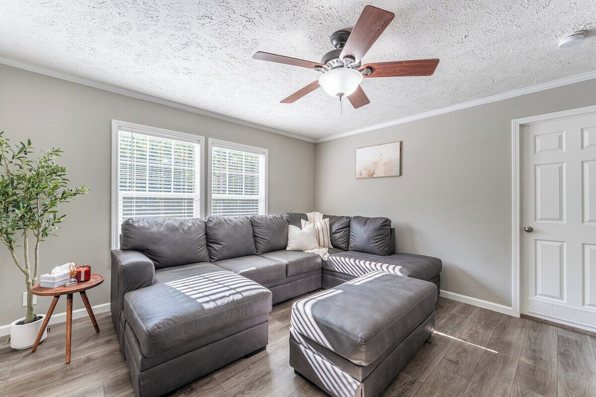 A living room with a sectional sofa, ottoman, ceiling fan, windows, and a door.