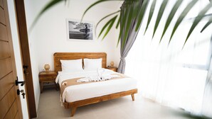 Un lit bien fait, avec du linge de lit blanc et une tête de lit en bois, dans une pièce lumineuse dotée d’une grande fenêtre et d’une plante.