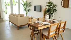 Un coin repas avec une table en bois dressée pour quatre personnes, entourée de chaises en osier. On aperçoit au fond un canapé beige, un téléviseur fixé au mur et plusieurs plantes en pot.