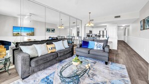 A spacious living room with two grey sofas, a glass coffee table, and a large mirror reflecting the kitchen area. - Majestic Sun 805A | Unobstructed Views, 8th Floor! | Close to Shops & Dining | Perfect Weekend Getaway Spot! (Miramar Beach)