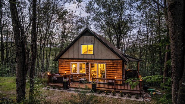 A cabin with a wooden exterior, a deck with outdoor seating, and a large window. - Finley Lodge | 5 Mins to Attitash | Game Room (Bartlett)