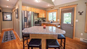 A kitchen with a granite island, stainless steel appliances, wooden cabinets, and a window with a view of greenery. - Finley Lodge | 5 Mins to Attitash | Game Room (Bartlett)