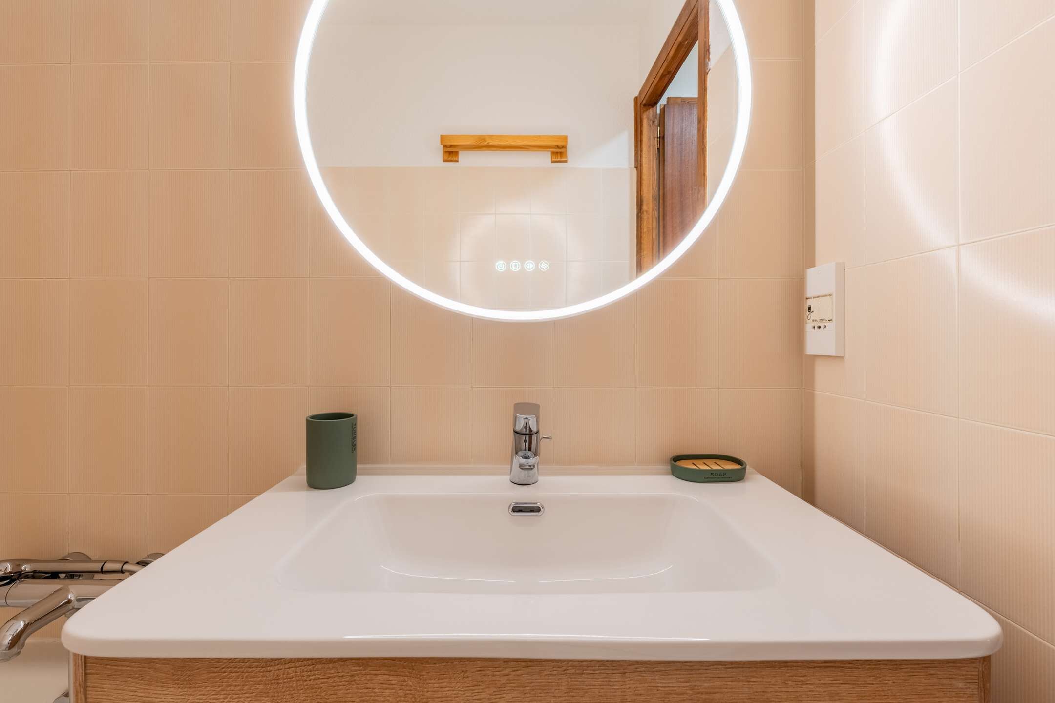 Ett badrum med ett vitt handfat, en spegel med en rund lampa, en trähylla och en toalett.