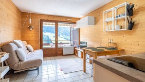 A cozy cabin with a sofa, a dining table, and a kitchen area with a hanging shelf. - Champel A : Appartement lumineux, panorama alpin (LE GRAND BORNAND)