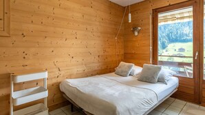 A wooden cabin bedroom with a bed, pillows, a white shelf unit, and a window with a view of a forest. - Champel A : Appartement lumineux, panorama alpin (LE GRAND BORNAND)
