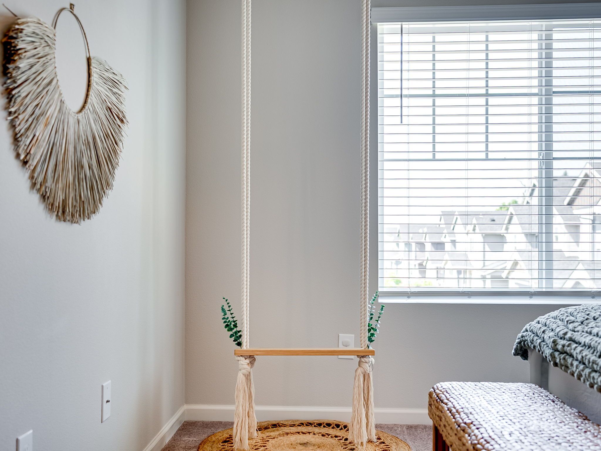 A room with a macrame wall hanging, a macrame swing, and a view through a window with blinds.