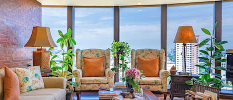 Un salón amplio con dos sillones estampados, un sofá, una mesa de centro con libros y un jarrón, y ventanas grandes con vistas a la ciudad.