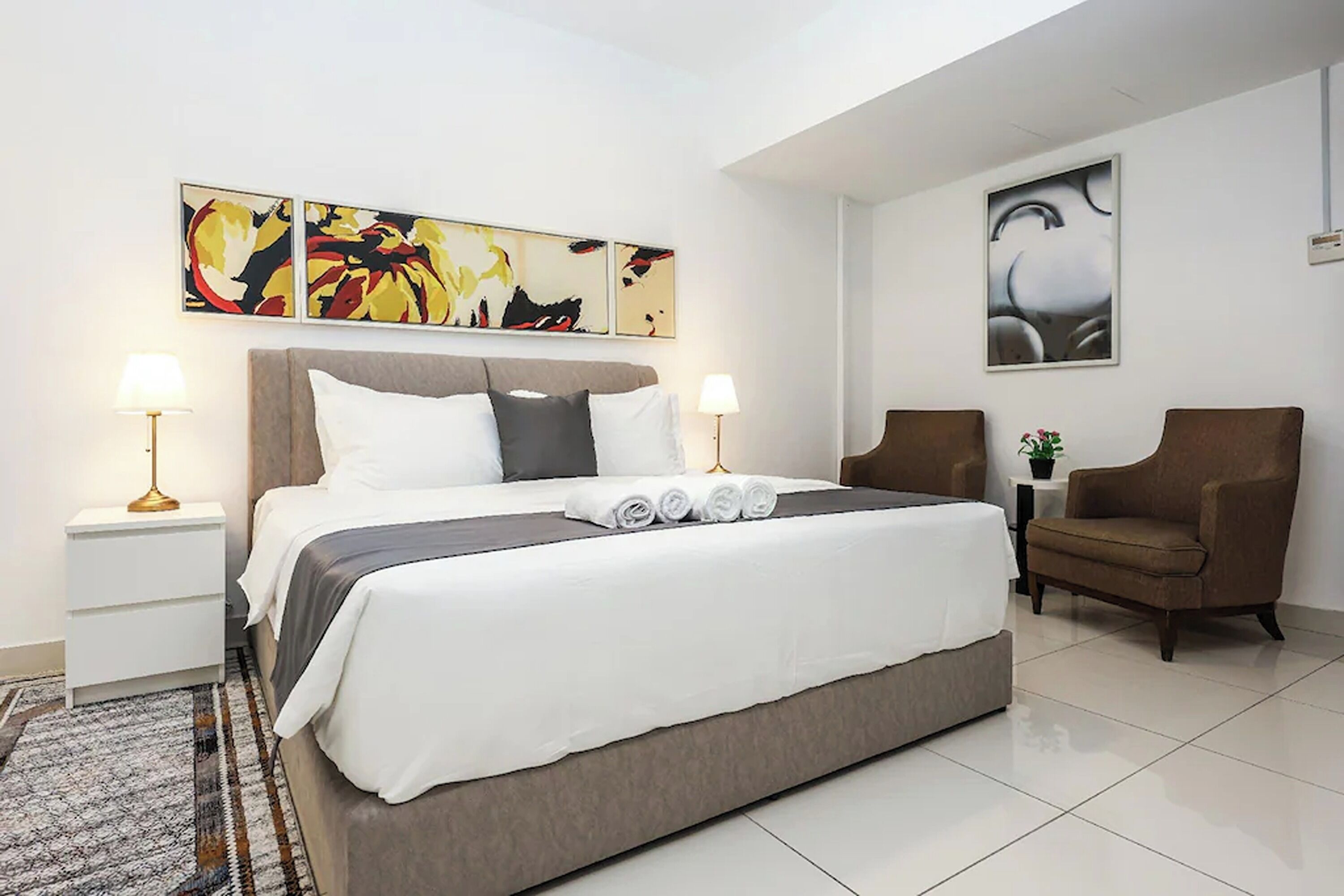 Una habitación de hotel moderna con una cama grande, dos mesitas de noche, una silla y arte abstracto en la pared.