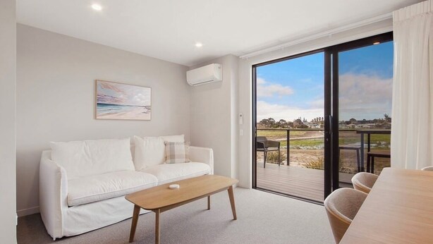 A modern living room with a white sofa, a wooden coffee table, and a large sliding glass door leading to an outdoor patio. - Relaxing Home in Waihi Beach (#B2) (Waihi Beach)