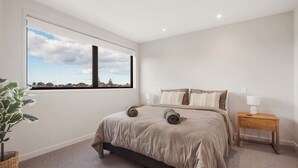A bedroom with a large window, a bed with a beige bedspread, two bedside tables with lamps, and a potted plant. - Relaxing Home in Waihi Beach (#B2) (Waihi Beach)