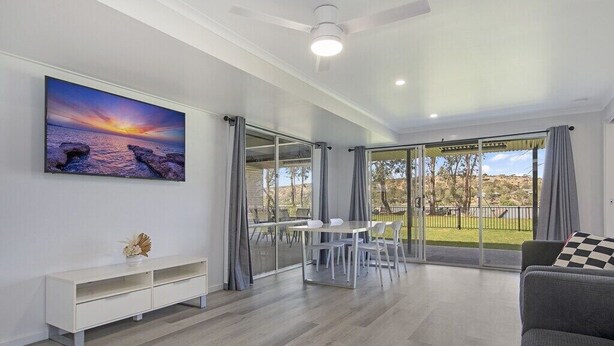 A spacious living area with a dining table, a sofa, a large painting, and a sliding glass door leading to an outdoor area. - Absolute Waterfront Bliss – Pet Friendly, Seperate living areas, Family Shack. (Pellaring Flat)