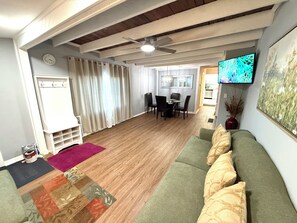 A living room with a green sofa, a dining area with chairs, a ceiling fan, and a television on the wall.