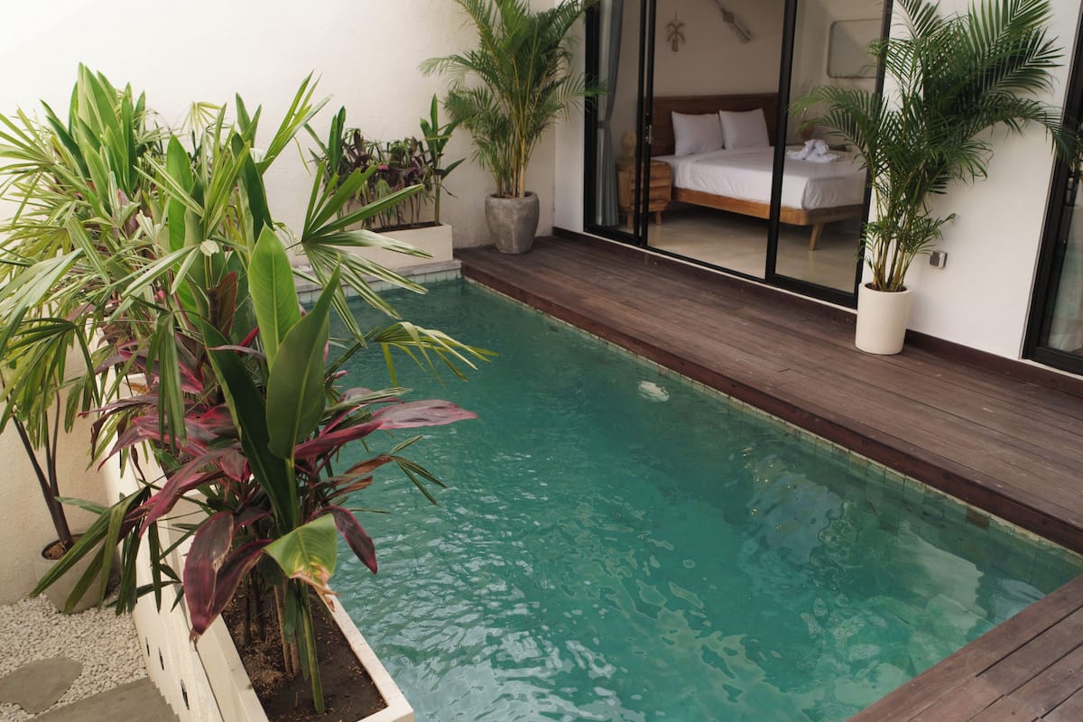 A modern pool area with a wooden deck, surrounded by potted plants and a glass door leading to a bedroom.