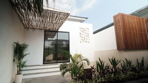 Een modern gebouw met een witte gevel, een glazen deur en een houten hek. Er zijn treden die naar de ingang leiden, en diverse planten zijn zichtbaar.