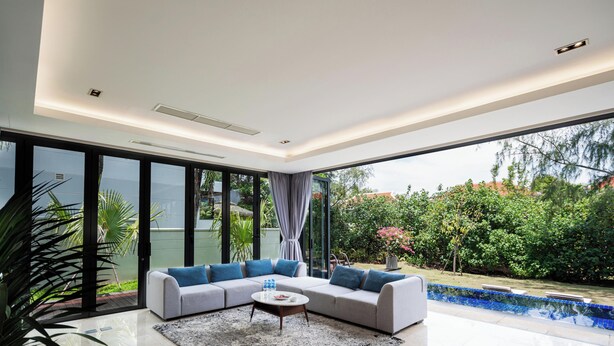 A modern living room with a sectional sofa, a coffee table, and a view of a swimming pool and greenery. - Danang private pool villa by Esea43 (Da Nang)