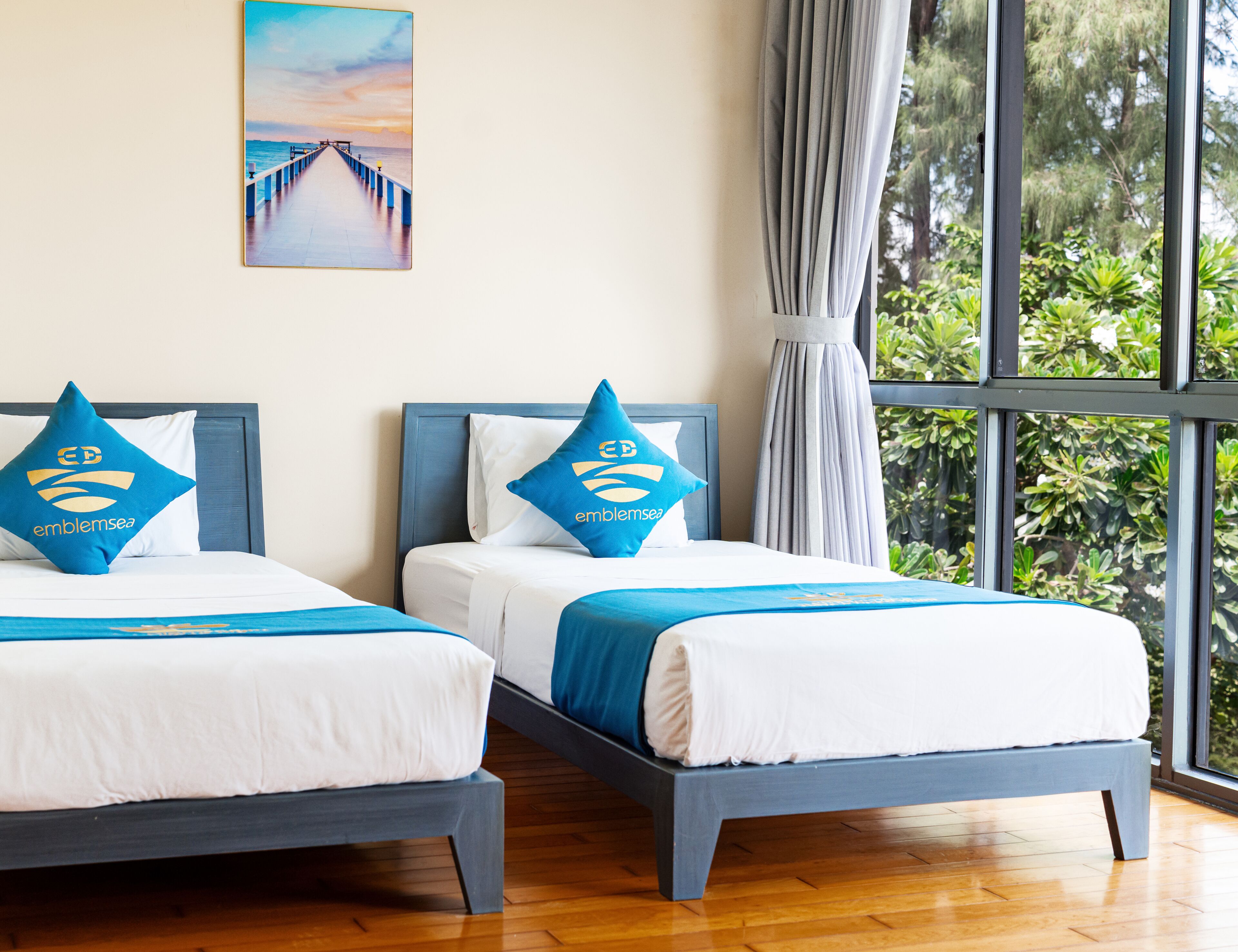 Een hotelkamer met twee bedden, elk voorzien van een blauw-witte dekbedovertrek en bijpassende kussens. Daarboven hangt een schilderij van een pier aan de muur.