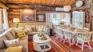 Interior acogedor de una cabaña con techo de madera, un comedor con sillas blancas, un sofá con cojines y una mesa de centro con objetos decorativos.
