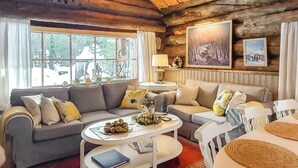 Un acogedor interior de cabaña de troncos con un sofá gris, una mesa redonda de centro y una mesa de comedor con individuales tejidos.
