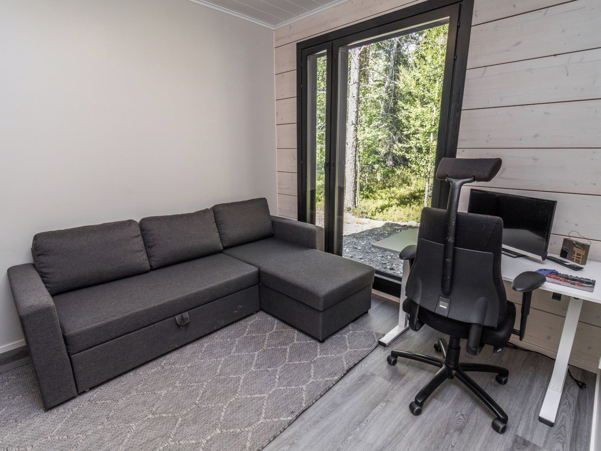 A modern room with a sectional sofa, a desk with a computer, and a view of a forest through a sliding door.