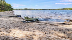 Una playa arenosa con un bote salvavidas y un bote pequeño sobre un remolque, un muelle de madera y una costa arbolada.