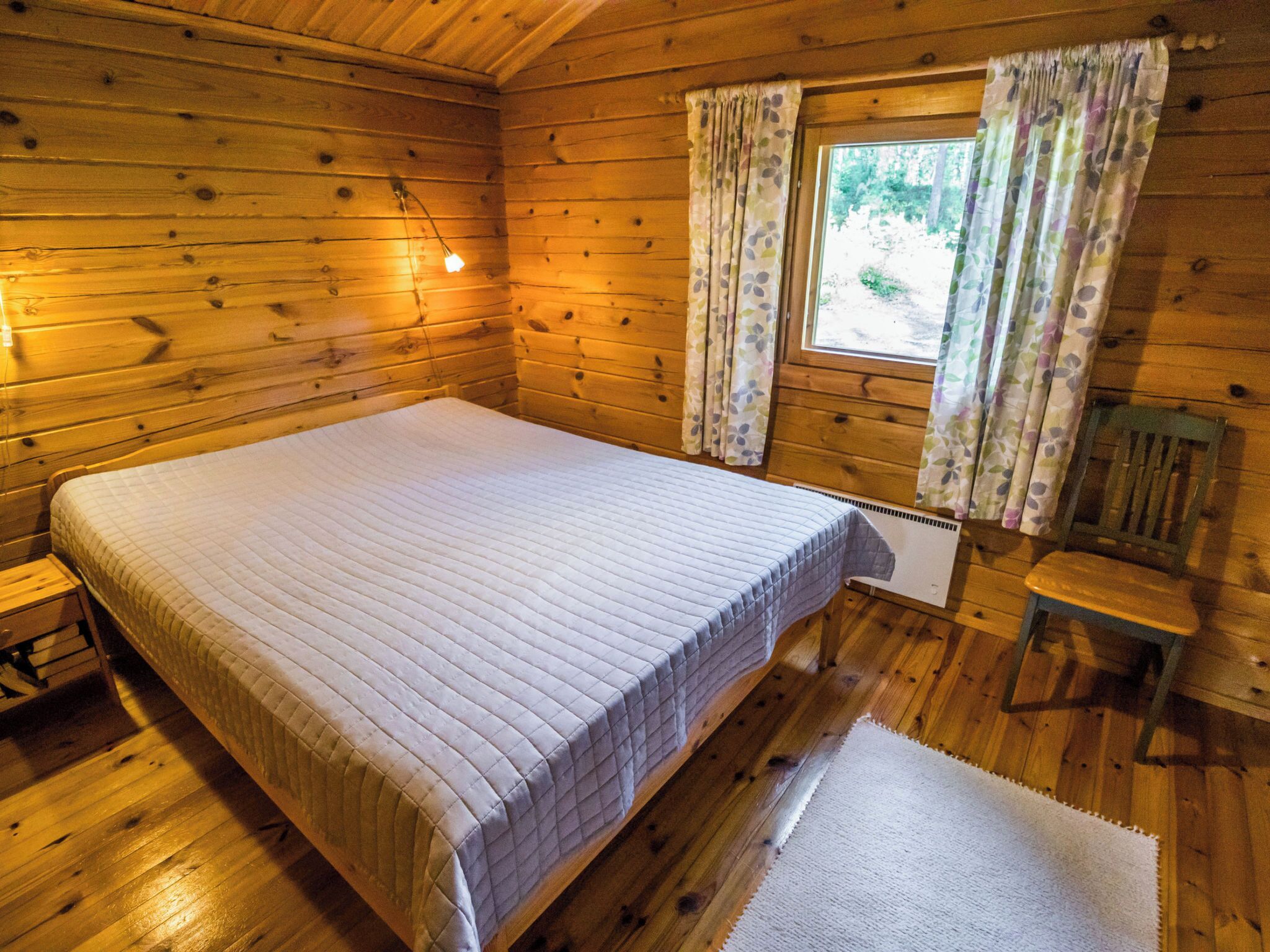A wooden cabin bedroom with a large bed, a nightstand, a chair, and a window with curtains.