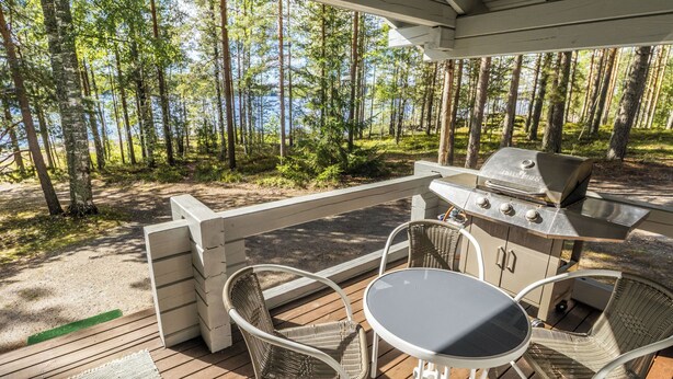 En utomhusveranda med en grill, flätade stolar och ett bord, med utsikt över en skog och en sjö.