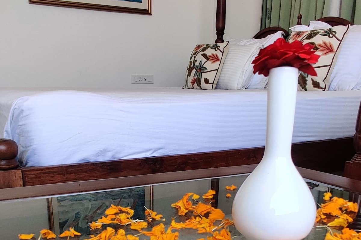 A room with a bed, a white vase with red flowers, and scattered orange petals on the floor.