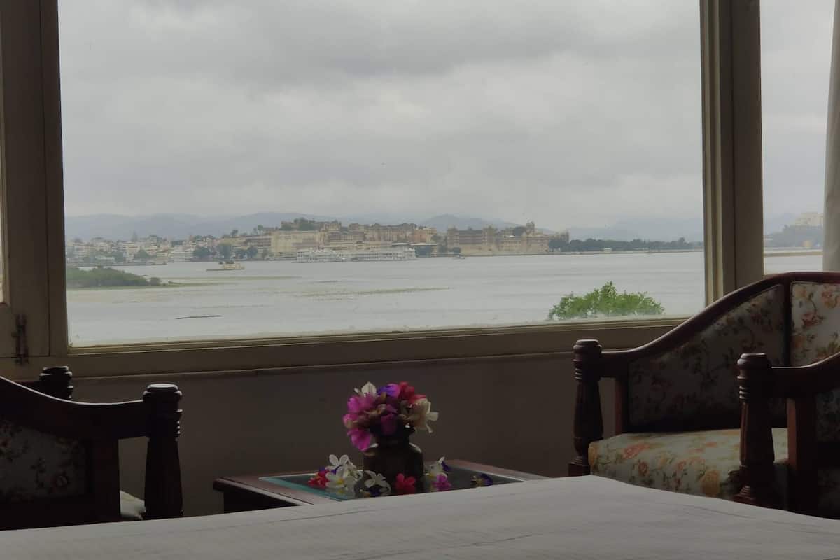 A room with a large window overlooking a river and distant buildings. There is a table with a vase of flowers and two chairs.