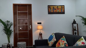A living room with a wooden door, a black sofa with colorful cushions, a wall-mounted Buddha head sculpture, a framed painting, and a floor lamp. - Cozy and Tranquil Nest ( With wifi and Homely Care ) (Greater Noida)